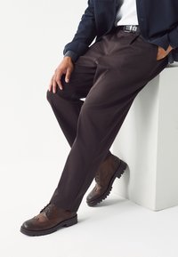 Dunkelbraune Hose mit geradem Schnitt, gefertigt aus glattem Stoff. Kombiniert mit braunen Lederschuhen mit dekorativer Brogue-Verzierung.