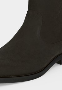 Gros plan sur l’avant et le côté d’une bottine en daim brun foncé avec un bout arrondi et un petit talon.