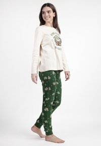 Top de manga larga color crema claro con gráfico. Pantalones de pijama verdes con personajes de dibujos animados y estrellas impresas. Tela suave, corte relajado.