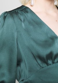 Robe en satin vert avec un décolleté en V et des manches bouffantes, présentant une taille cintrée et un doux éclat. Une boucle d'oreille en perle pendentive ajoute une touche de détail.