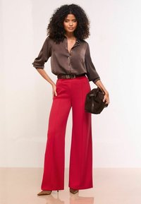 Blusa in satin marrone con bottoni, abbinata a un paio di pantaloni rossi a vita alta e gamba larga e una borsa in suede marrone. Tacchi beige a punta.