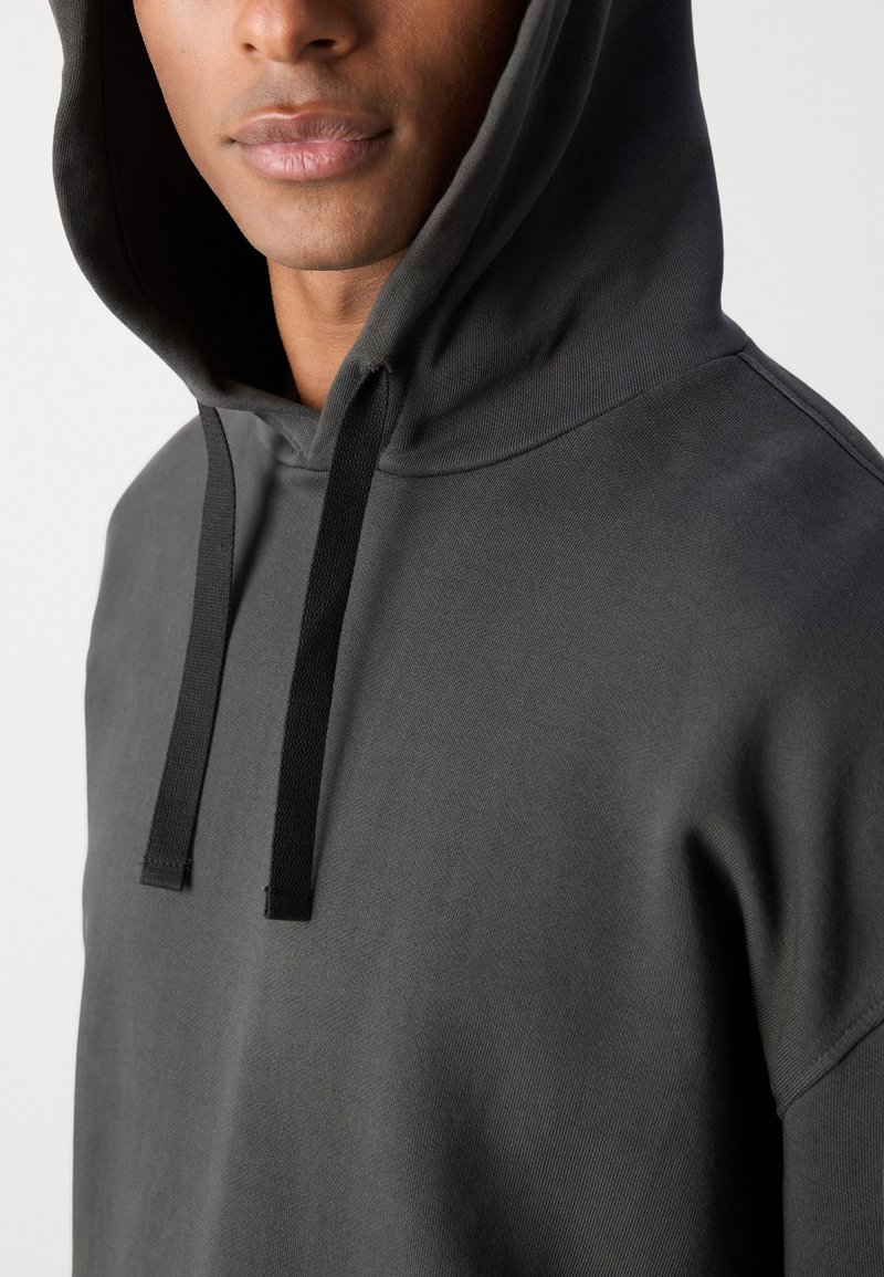 Donkergrijze hoodie van zacht materiaal, met een grote capuchon, zwarte trekkoorden en een relaxed fit. Beperkte zichtbare stiksel details.