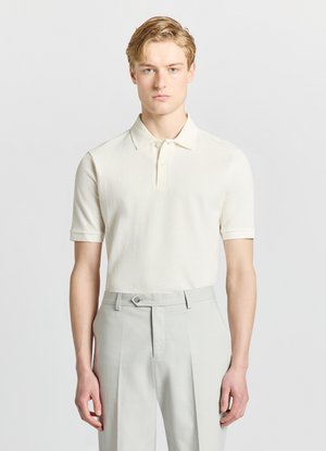 Giovane uomo che indossa una polo bianca a maniche corte con texture infilata in pantaloni grigio chiaro su misura, in piedi davanti a uno sfondo chiaro e uniforme.