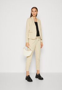 Blazer beige ajusté sur un top noir, associé à un pantalon beige et des mocassins noirs en cuir épais. Tenant un sac à main en forme de croissant blanc.
