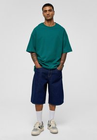 Overmaatse teal T-shirt, blauwe denimshorts, witte sokken en sneakers. Heeft tatoeages op de armen, met een casual pasvorm en een relaxte stijl.