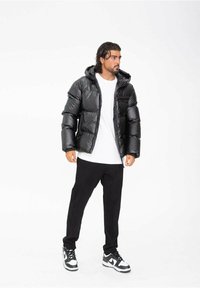 Doudoune noire avec une finition brillante et une capuche, dotée d'une fermeture éclair et de poches latérales, portée par-dessus un t-shirt blanc et un pantalon noir.