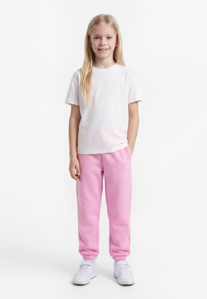 Jeune fille aux longs cheveux blonds portant un t-shirt blanc, un pantalon de survêtement rose et des baskets blanches, debout sur un fond blanc uni.