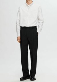 Man klädd i svarta byxor, svarta skor och en vit långärmad skjorta med ljusa vertikala ränder, stående med händerna i fickorna.