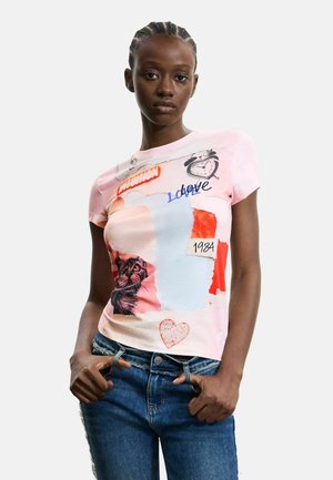 Ung kvinde med flettet hår iført en pink T-shirt med grafik af en kat, ur, "Love", "1984" og hjerter, kombineret med blå jeans.