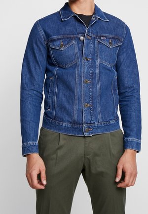 Mann trägt eine blaue Jeansjacke mit Knopfleiste und Brusttaschen, kombiniert mit olivgrünen Hosen vor einfachem Hintergrund.