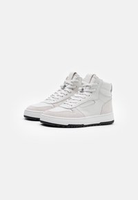 Sneaker alte bianche con accenti in suede, suole nere spesse e lacci bianchi, disposte affiancate su uno sfondo bianco semplice.