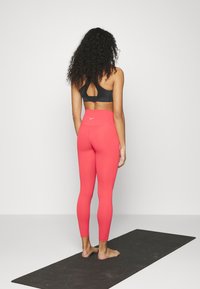 Czarny sports bra z kluczem z tyłu, w połączeniu z obcisłymi, wysokimi legginsami w kolorze koralowym. Stoi na ciemnej macie do jogi, boso.