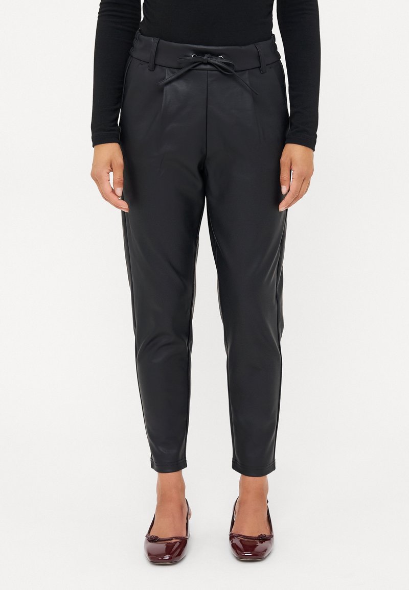 Personne portant un pantalon noir ajusté avec une taille à cordon et des talons en cuir verni bordeaux, debout devant un fond blanc.