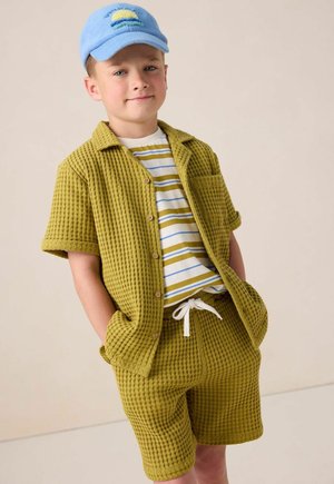 Ensemble en vert texturé comprenant une chemise et un short avec une poche. T-shirt blanc à rayures en dessous. Casquette bleu clair avec un motif de soleil.