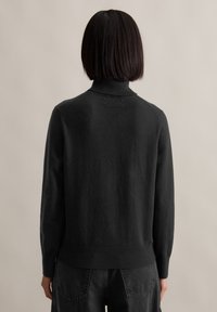 Rövid sötét hajú személy fekete garbót és fekete farmernadrágot visel, háttal állva egy sima bézs háttér előtt.