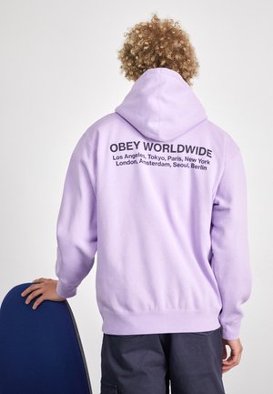Osoba z kręconymi włosami w jasnofioletowej bluzie z kapturem z napisem „OBEY WORLDWIDE” i listą głównych miast na plecach, opierająca rękę na niebieskim krześle.