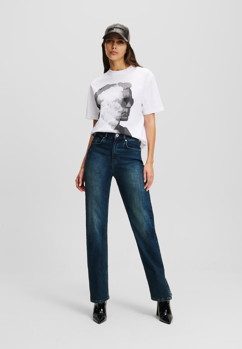 White graphic t-shirt featuring a monochrome face design, paired with dark blue straight-leg jeans and black pointed ankle boots.