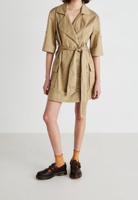 Khaki krótka sukienka trench z paskiem, dużymi guzikami i dwiema przednimi kieszeniami. W połączeniu z brązowymi butami i pomarańczowymi skarpetkami w prążki.