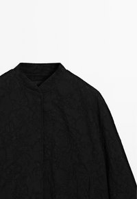 Veste noire avec un motif floral subtil, avec un col haut et une fermeture frontale cachée sur un fond uni.