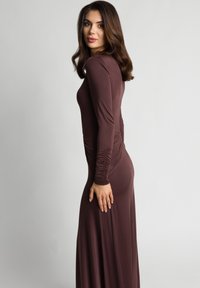 Długa brązowa sukienka z długim rękawem, z marszczonymi detalami przy rękawach i po bokach, wykonana z gładkiego, elastycznego materiału.