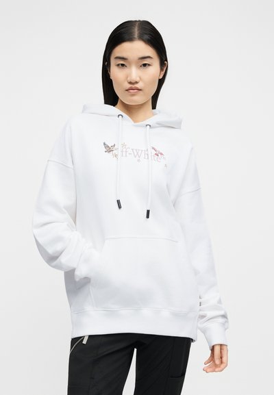 Νεαρή γυναίκα που φοράει λευκό φούτερ Off-White με σχέδια πουλιών και αστεριών, μαύρο παντελόνι, στέκεται μπροστά σε απλό φωτεινό φόντο.