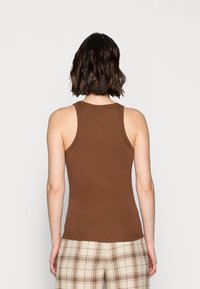 Femme aux cheveux courts et foncés, portant un haut marron sans manches et un pantalon à carreaux beige, debout de dos sur un fond uni.