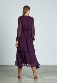 Vestido largo de maxi en tono púrpura con mangas largas, puños con volantes y cintura ceñida, hecho de una tela ligera y transparente. Botas marrones hasta la rodilla.