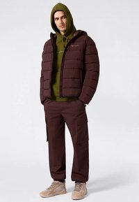 Braune Steppjacke mit vertikalen Nähten, darunter olivgrüner Kapuzenpullover, und braune Cargohosen. Beigefarbene Sneakers vervollständigen das Outfit.