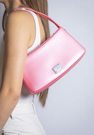 Woman with long brown hair wearing white top and pants, carrying smooth pink handbag with silver rectangular logo plaque on shoulder.