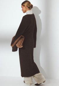 Long brown ribbed knit cardigan, oversized fit, open front, with wide sleeves. Paired with light-colored wide-leg pants and a beige clutch.