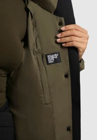 Olijfgroene pufferjack binnenkant met zwarte accenten, voorzien van een merklabel. Glad fabric, knoopsluitingen en een subtiele glans.