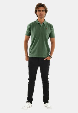 Grünes Poloshirt mit kurzen Ärmeln und kleinem Logo. Kombiniert mit schwarzer Hose und weißen Turnschuhen. Model steht mit entspannten Armen.