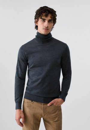 Giovane uomo con capelli scuri e ricci indossa un maglione dolcevita grigio scuro e pantaloni marrone chiaro, in piedi davanti a uno sfondo chiaro e semplice.