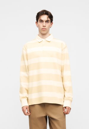 Fiatal férfi bézs-fehér csíkos hosszú ujjú galléros ingben és világosbarna nadrágban, egyszínű világos háttér előtt áll.