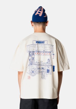 Persoon draagt een crèmekleurig T-shirt met blauwe lijntekening van een noedelwagen en de tekst "WOODBIRD" en "NOODLES" op de rug, gecombineerd met een blauwe muts.