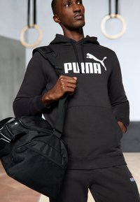 Schwarzer Kapuzenpullover mit "PUMA"-Logo, kombiniert mit einer schwarzen Sporttasche mit Netzdetails. Glattes Material, entspannte Passform und verstellbare Riemen.