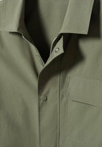 Giacca verde oliva con chiusura a zip, colletto a punta e tasca sul petto. Realizzata in tessuto morbido con cuciture a vista.