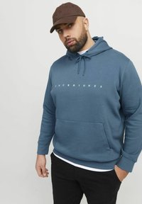 Hoodie in blauw, met een frontzak en witte tekst "JACK & JONES." Gekleed met zwarte broek en een bruine pet.