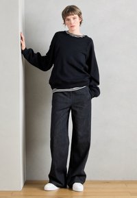 Melns pārliels džemperis ar garām piedurknēm pār melnu un baltu svītrainu topu, kopā ar platajam kāju tumšajiem džinsa biksēm un baltām sporta kurpēm.