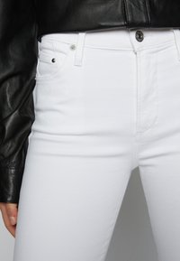 Vaqueros de denim blanco con un corte ajustado, que cuentan con herrajes plateados, diseño de cinco bolsillos y una superficie texturizada. Combinados con una chaqueta de cuero negra.