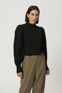 Blusa nera plissettata con colletto alto e chiusura a bottone; abbinata a pantaloni beige a righe. Maniche lunghe con dettaglio arricciato.