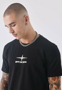 Zwarte T-shirt met korte mouwen, featuring een witte "PXP" logo en ster ontwerp aan de voorkant. Gladde stof met een aangesloten silhouet. Zilveren ketting.