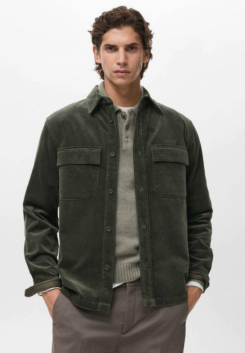 Joven vestido con una chaqueta de botones de pana verde oscura sobre un suéter beige y pantalones marrones, de pie con las manos en los bolsillos.