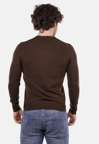 Uomo con capelli ricci che indossa un maglione marrone aderente e jeans blu, in piedi di spalle su uno sfondo bianco uniforme.