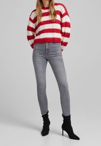 Červeno-bílý pruhovaný pletený svetr, šedé skinny džíny s dírou, černé vysoké slouchy kotníkové boty. Jednoduché pozadí.