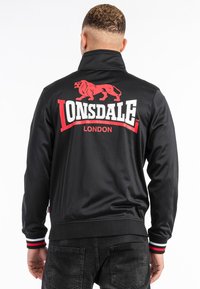 Svart jacka med hög krage, prydd med en röd lejonlogotyp och texten "LONSDALE LONDON" i vitt. Ribbad mudd med röda och vita detaljer.