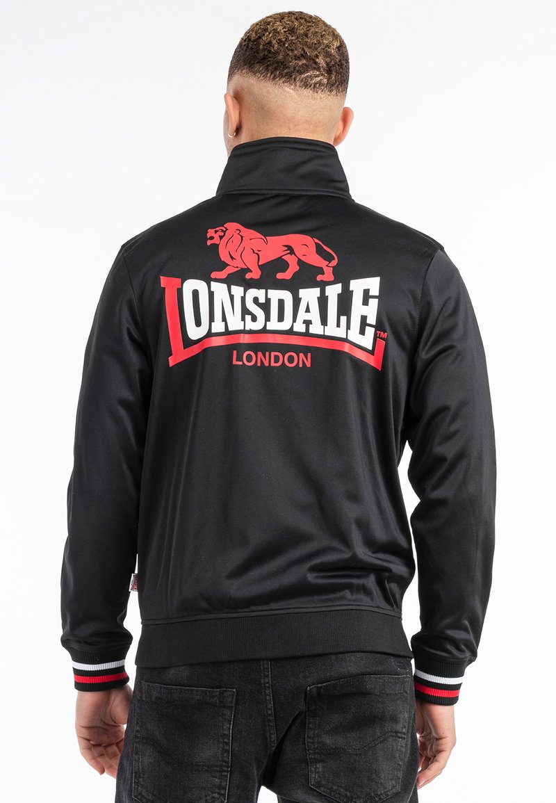 Svart jacka med hög krage, prydd med en röd lejonlogotyp och texten "LONSDALE LONDON" i vitt. Ribbad mudd med röda och vita detaljer.