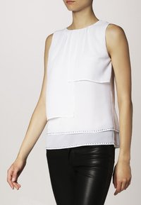 Top bianco senza maniche con design a strati, scollatura plissettata e borchie decorative lungo l'orlo, abbinato a pantaloni neri aderenti.
