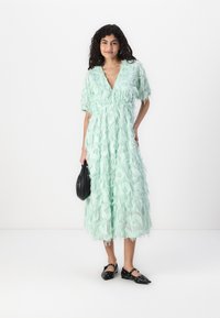 Mint green dress made of textured fabric with layered fringe detailing, short sleeves, and a deep V-neck. Worn with black flats and a bag.