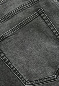 Mustan denimin kangas, jossa näkyy takatasku selkeinä ompeluyksityiskohtina. Tekstuurissa on hieman kulunutta ilmettä ja sileä pinta.